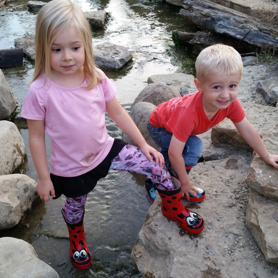 A young blonde girl and a young blonde boy stand on rocks in the stream of the Nature PlayScape